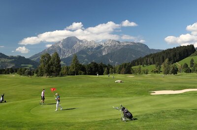 Golf Club Urslautal Golfplatz mit Spielern auf dem Green vor einer alpinen Bergkulisse unter blauem Himmel.