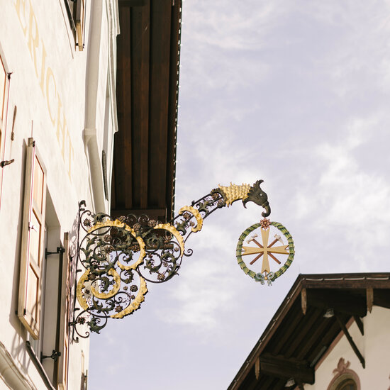 Ein kunstvoller, geschmückter Wirtshausschild hängt an einer historischen Fassade in Kitzbühel, unter einem blauen Himmel.
