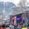 Eine große Menschenmenge versammelt sich vor einer Bühne, während ein Sänger mit Mikrofon auftritt, umgeben von schneebedeckten Bergen.