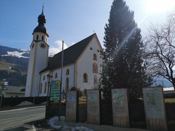 Weiße Kirche mit Zwiebelturm neben Informationstafeln an einer Straße vor Bergen.