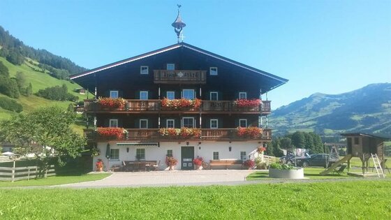 Sommer Aussen am Angererhof Traditionelles Alpenhaus mit Blumenbalkonen vor bewaldeten Bergen unter blauem Himmel.