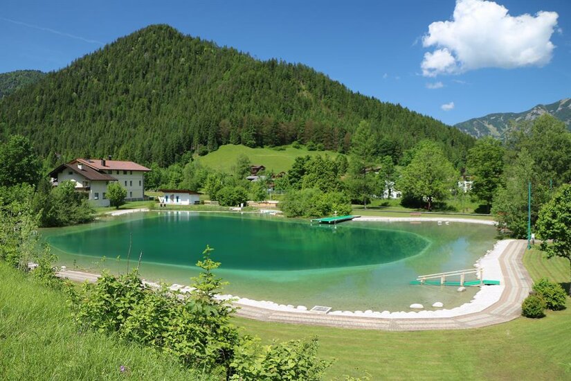 Ein ruhiger See umgeben von grünen Hügeln und dichten Wäldern. Im Hintergrund sind ein paar Häuser und ein klarer blauer Himmel sichtbar.