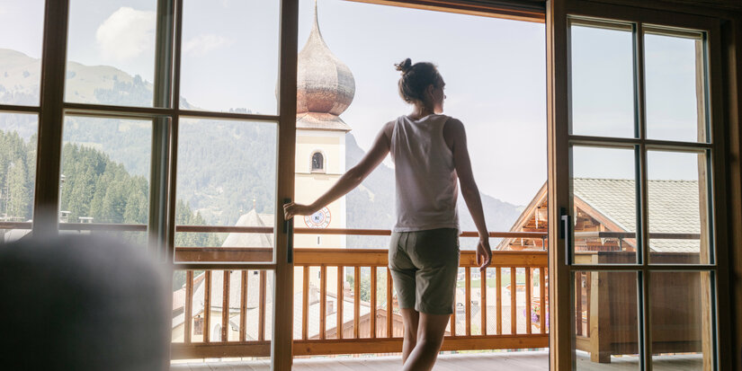 Durch das Fenster sieht man eine Person, die die ruhige Berglandschaft und den historischen Kirchturm bewundert.