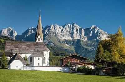 Ortsansicht Reith mit Kaisergebirge Weiße Kirche mit spitzem Turm vor einem massiven Gebirgszug unter blauem Himmel.