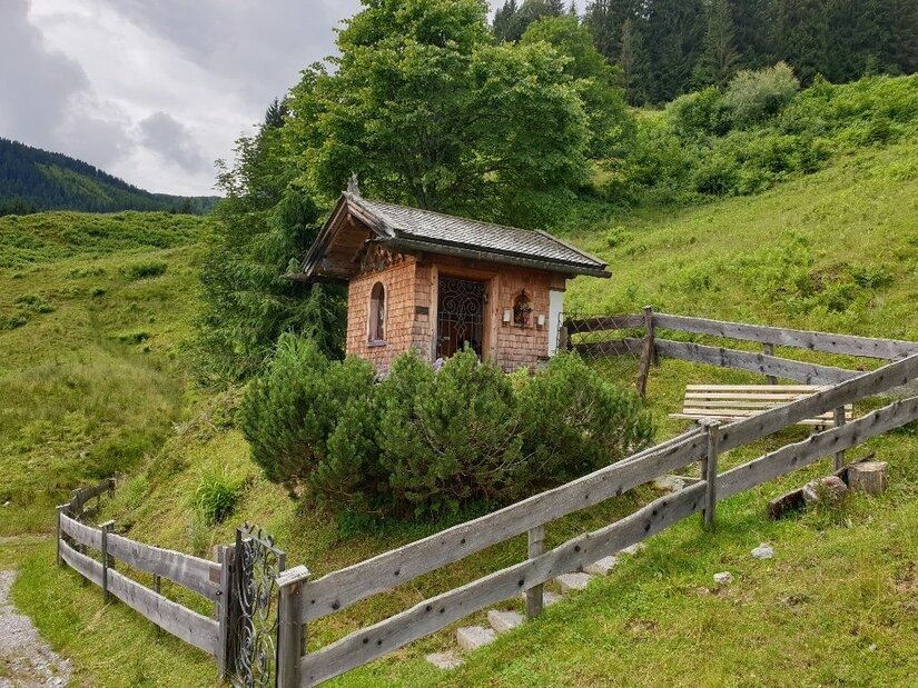 Kapelle bei der Kinzlingalm Kleine Holzkapelle mit Schindelfassade auf einer grünen Almwiese hinter einem Holzzaun.