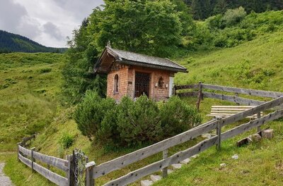 Kleine Holzkapelle mit Schindelfassade auf einer grünen Almwiese hinter einem Holzzaun.