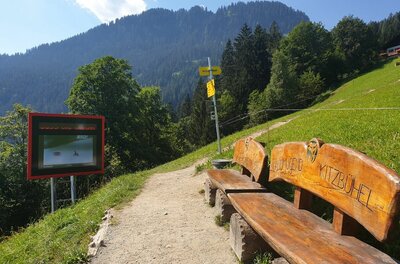 Wanderweg mit Holzbank, Infotafel und Wegweisern vor bewaldetem Berghang.