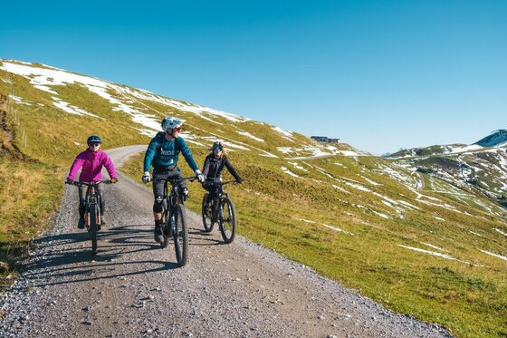 Mountainbiken in Kitzbühel Drei Mountainbiker fahren auf einem Schotterweg in einer alpinen Graslandschaft mit Schneeresten unter blauem Himmel.