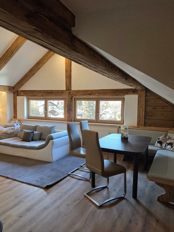 Ein modernes Wohnzimmer mit Holzbalken und großen Fenstern. Es gibt eine gemütliche Sofagarnitur und einen Esstisch mit Stühlen.