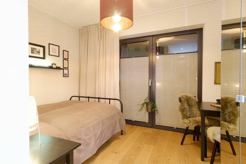 Schlafbereich im Apartment Modernes Schlafzimmer mit Metallbett, Holzboden und einem kleinen Tisch mit Fellstühlen vor einem Fenster.