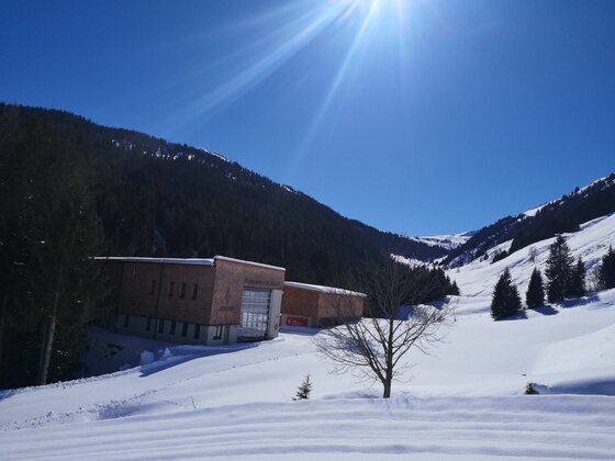 Bruggeralm im Winter Modernes Holzgebäude in einer verschneiten Gebirgslandschaft unter blauem Himmel mit Sonneneinstrahlung.