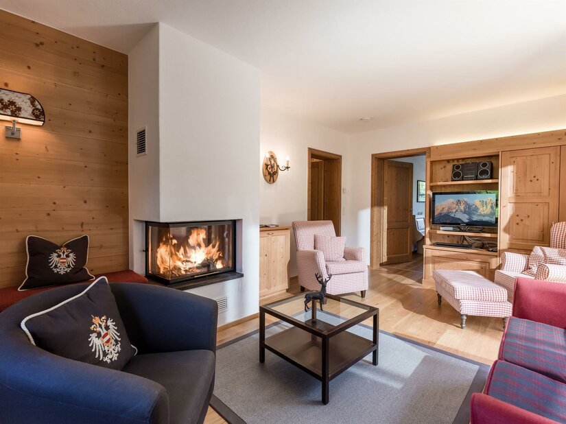 Wohnbereich Appartement Hahnenkamm Wohnzimmer mit brennendem Kamin, Sesseln, Couchtisch und Fernseher in alpiner Holzoptik.