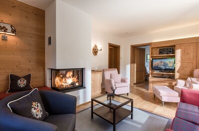 Wohnbereich Appartement Hahnenkamm Wohnzimmer mit brennendem Kamin, Sesseln, Couchtisch und Fernseher in alpiner Holzoptik.