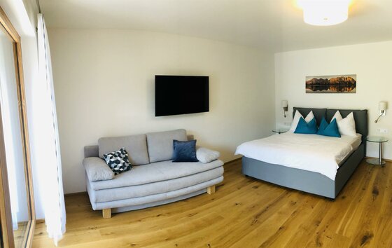 Modernes Schlafzimmer mit Doppelbett, grauem Sofa, Flachbildfernseher und Holzboden.