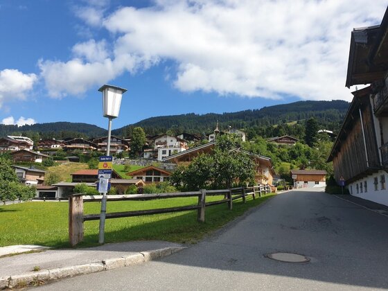 Asphaltierte Straße in einer Siedlung mit Holzhäusern vor bewaldeten Bergen unter blauem Himmel.
