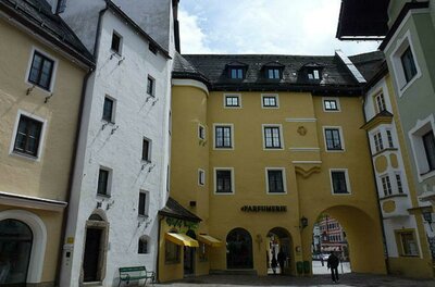 Ein malerischer Platz mit bunten Gebäuden und einem gelben Haus mit einem Tor. Die Architektur zeigt einen typischen, charmanten Stil.