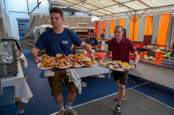 Feuerwehrfest Zwei Männer tragen mehrere Teller mit Essen auf einem Fest. Im Hintergrund sind weitere Personen und einen Imbissbereich zu sehen.