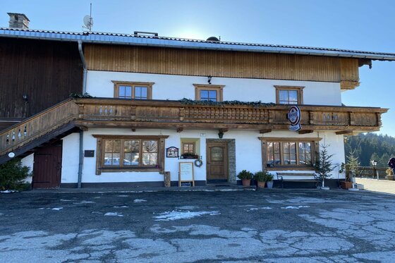 Vorderansicht eines zweistöckigen Gasthofs aus Holz und weißem Putz mit Balkon und Parkplatz im Winter.