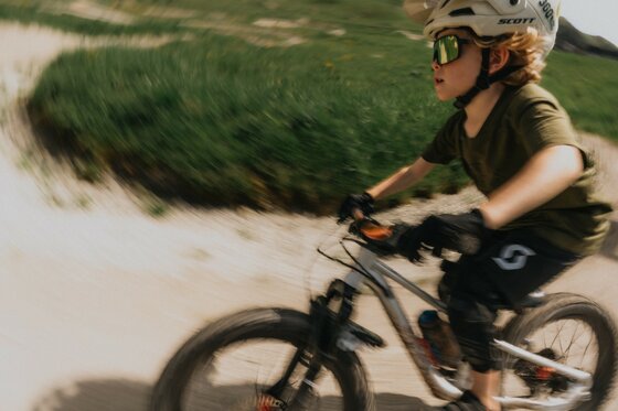 Ein Kind mit Helm und Sportbrille fährt Mountainbike auf einem Schotterweg in einer Kurve.