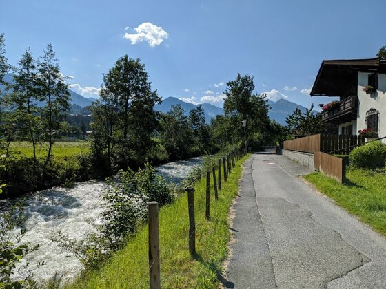 Asphaltierter Weg neben einem fließenden Bach mit Holzzaun, Bäumen und Bergen im Hintergrund.