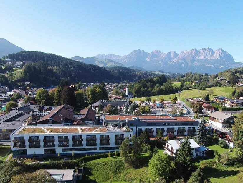 Luftaufnahme eines modernen Klinikgebäudes in Kitzbühel vor bewaldeten Hügeln und dem Wilden Kaiser.