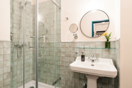 Badezimmer im HENRI Country House Modernes Badezimmer mit Glasdusche, weißem Standwaschbecken und rundem Spiegel vor hellgrünen Fliesen.