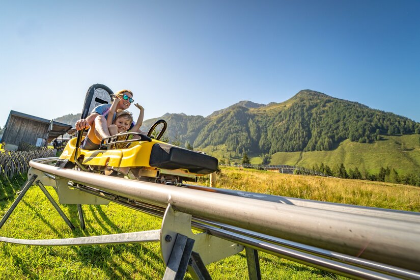 Timoks Coaster Sommer Berge Eine aufregende Sommerrodelbahn in den Bergen. Zwei Personen genießen die Fahrt bei schönem Wetter und klarem Himmel.