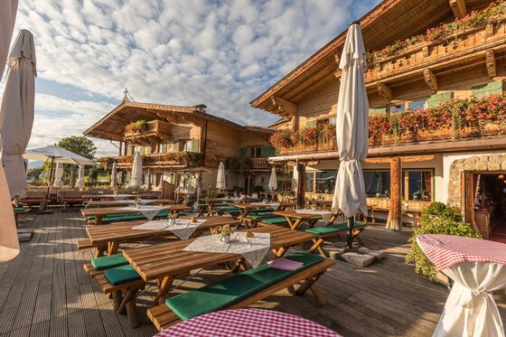 Terrasse der Sonnbergstuben Sonnenterrasse einer Holzhütte mit Holztischen, Bänken und Sonnenschirmen vor einem Alpenpanorama.