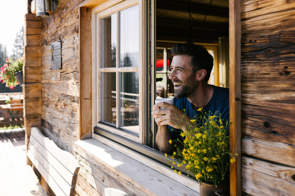 Der Mann am Fenster strahlt Freude aus, während er eine Tasse hält und die Natur von seiner Hütte aus betrachtet.