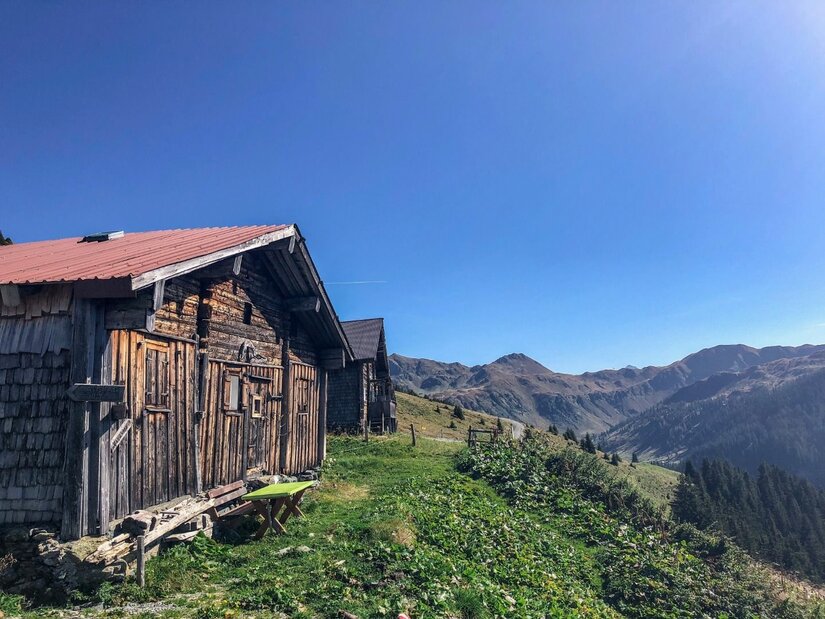 Almhütten am Schützkogel Alpinsteig Rustikale Holzhütten auf einer grünen Almwiese vor einem Gebirgspanorama unter blauem Himmel.
