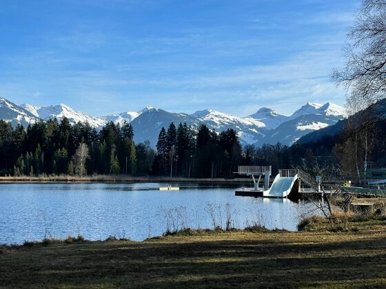Freibad am Schwarzsee Blick über einen See auf eine Badeanlage mit Rutsche und Sprungturm vor verschneiten Berggipfeln.