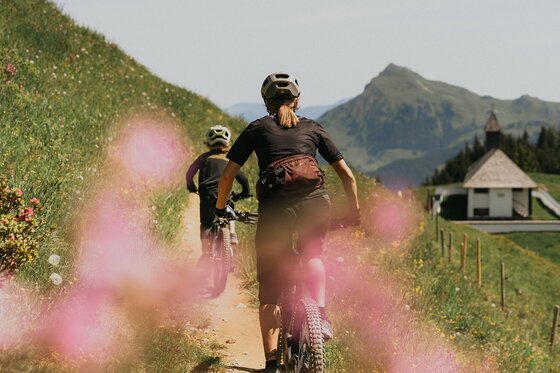 Hahnenkamm Trail mit Bergpanorama Zwei Mountainbiker fahren auf einem schmalen Pfad an einem Grashang entlang, im Hintergrund Berge und eine Kapelle.