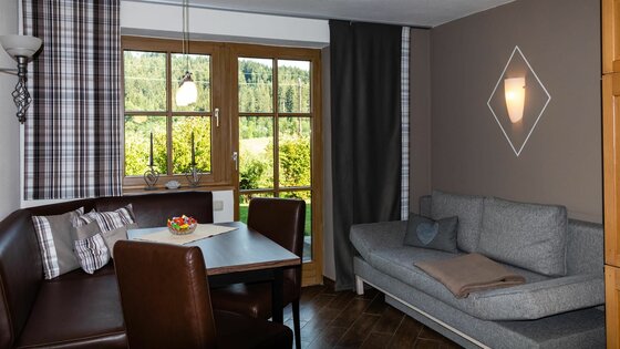Wohnbereich mit Eckbank, Holztisch und grauem Sofa vor einem Fenster mit Blick ins Grüne.