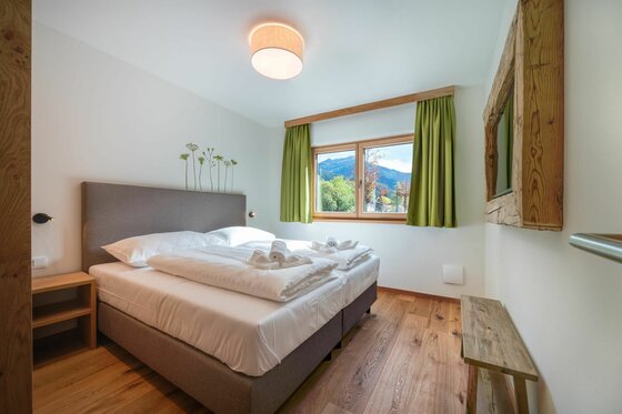 Schlafzimmer Wagstätt Residenz Jochberg Modernes Doppelzimmer mit Parkettboden, grauem Polsterbett und Fenster mit Bergblick.