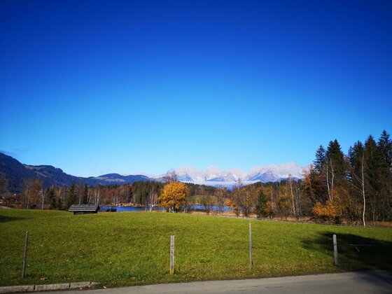 Wiese am See mit Bergblick Grüne Wiese mit Holzhütte vor einem See und herbstlichen Bäumen unter blauem Himmel mit Bergblick.