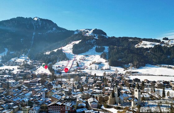 Hahnenkamm und Kitzbühel Luftaufnahme eines verschneiten Alpendorfes mit Blick auf eine Skipiste am bewaldeten Berghang.