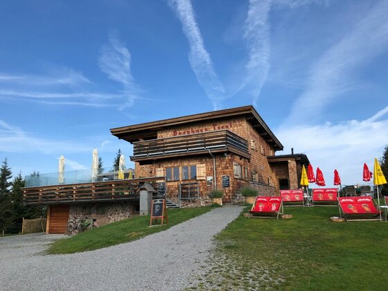 Holzgebäude mit Terrasse und roten Liegestühlen auf einer Wiese unter blauem Himmel.