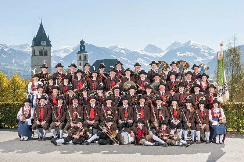Stadtmusik Kitzbühel Eine große Musikgruppe in traditionellen Trachten posiert vor einer malerischen Berglandschaft. Im Hintergrund sind historische Gebäude und schneebedeckte Berge zu sehen.