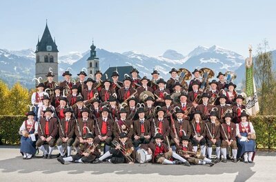 Eine große Musikgruppe in traditionellen Trachten posiert vor einer malerischen Berglandschaft. Im Hintergrund sind historische Gebäude und schneebedeckte Berge zu sehen.