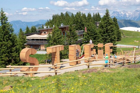 Spielplatz bei der Almhütte Große Holzbuchstaben mit dem Schriftzug STREIF auf einem Spielplatz vor einer Almhütte und Nadelbäumen.