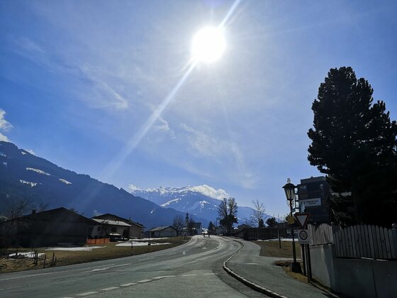 Ortsansicht Bärenbichl mit Bergpanorama Asphaltierte Straße in einer Siedlung mit Blick auf schneebedeckte Berge unter strahlender Sonne.
