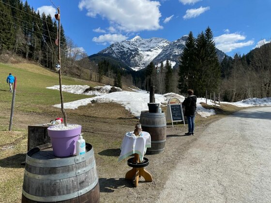 Wegstation bei der Burgeralm Holzfässer und ein Hinweisschild am Wegrand vor schneebedeckten Bergen.