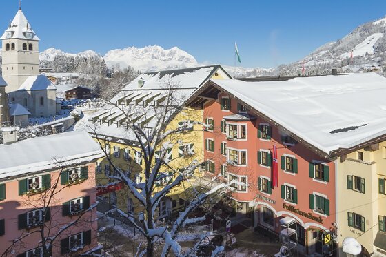 Verschneite Häuserzeile in Kitzbühel mit Kirchturm und schneebedeckten Bergen im Hintergrund.