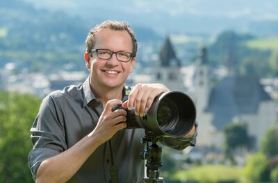 Ein lächelnder Fotograf mit einer Kamera steht im Freien. Im Hintergrund sind unscharfe Gebäude und eine grüne Landschaft zu sehen.
