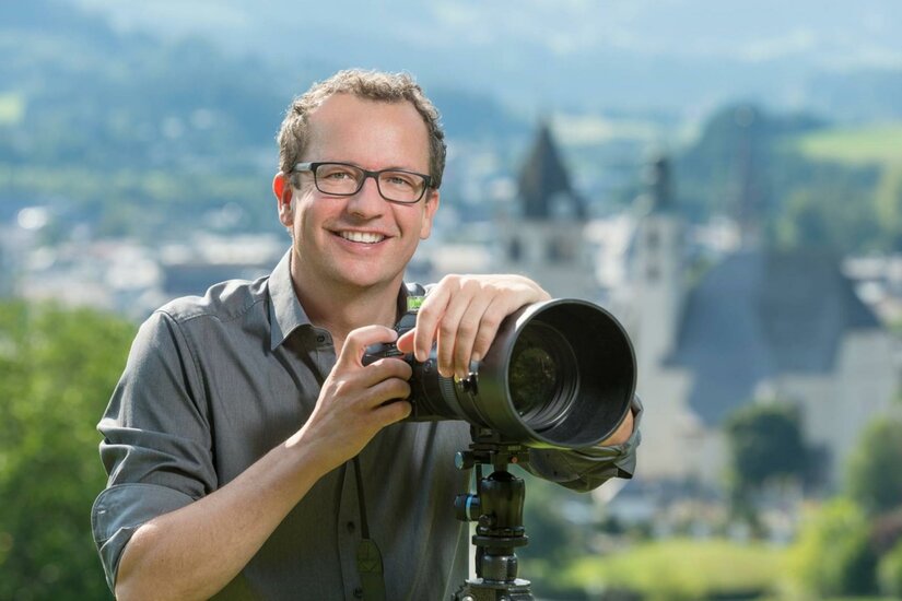 Markus Mitterer Fotografie Ein lächelnder Fotograf mit einer Kamera steht im Freien. Im Hintergrund sind unscharfe Gebäude und eine grüne Landschaft zu sehen.