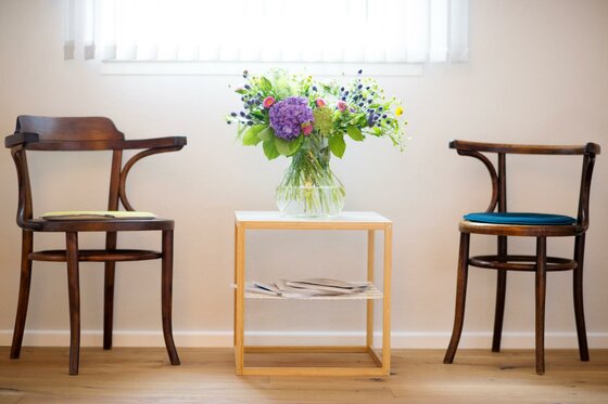 Zwei Holzstühle stehen neben einem kleinen Tisch. Auf dem Tisch steht eine Vase mit bunten Blumen.