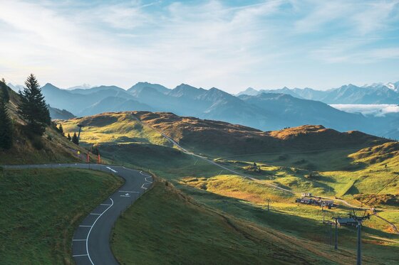 Asphaltierte Bergstraße führt in Kurven durch grüne Almwiesen vor einer Bergkette unter blauem Himmel.