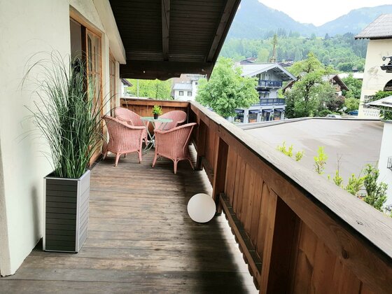 Balkon der Belle Kitz Holzbalkon mit rosa Korbsesseln, einem kleinen Tisch und Blick auf alpine Gebäude und bewaldete Berge.