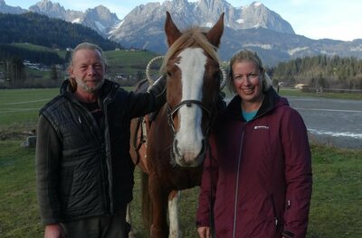 Ein Mann und eine Frau stehen neben einem braunen Pferd vor einer Bergkulisse.