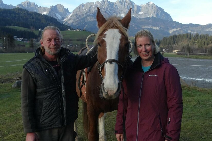 Ein Mann und eine Frau stehen neben einem braunen Pferd vor einer Bergkulisse.